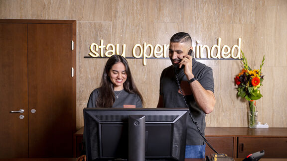 Magdas HOTEL c Karo Pernegger 59 Zwei fröhliche Mitarbeiter:innen stehen an der Rezeption von magdas HOTEL. An der Wand ist ein Schriftzug mit den Worten "stay open-minded" zu sehen.